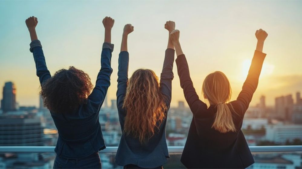 Victory! Three women stand in the sunrise, their backs to the viewer. Their fists are raised over their heads. Victory!